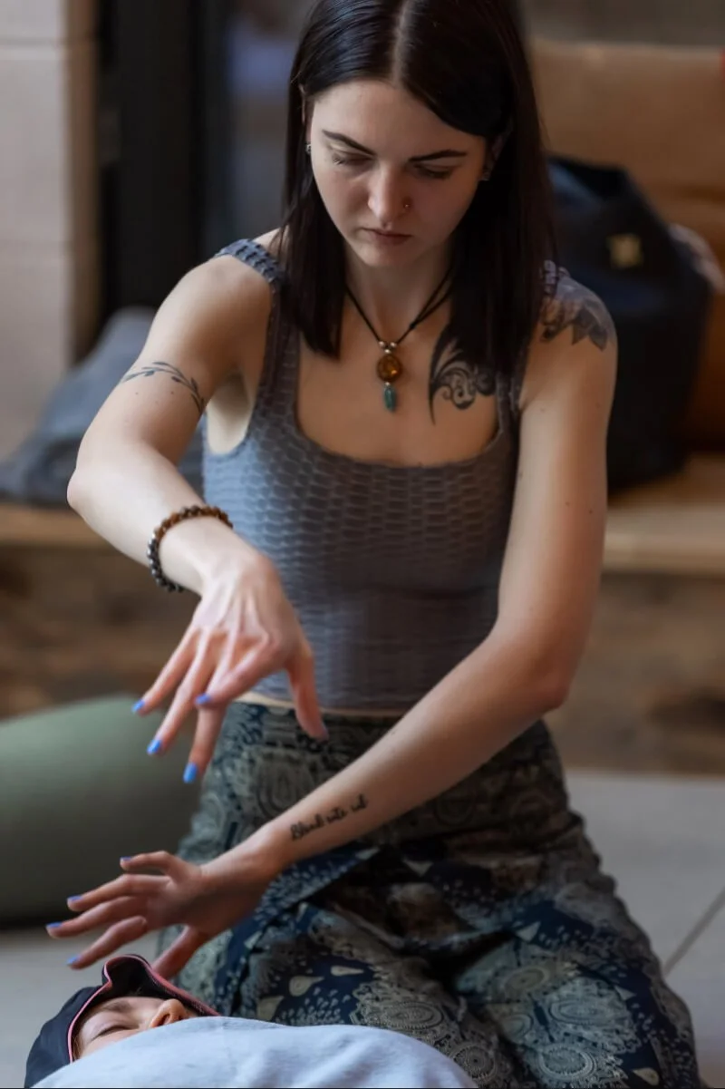 A woman with dark hair, wearing a sleeveless top, appears to be performing a healing session on a person laying down. She has tattoos on her arms, wears jewellery including a necklace with a pendant, and is focused on the task.