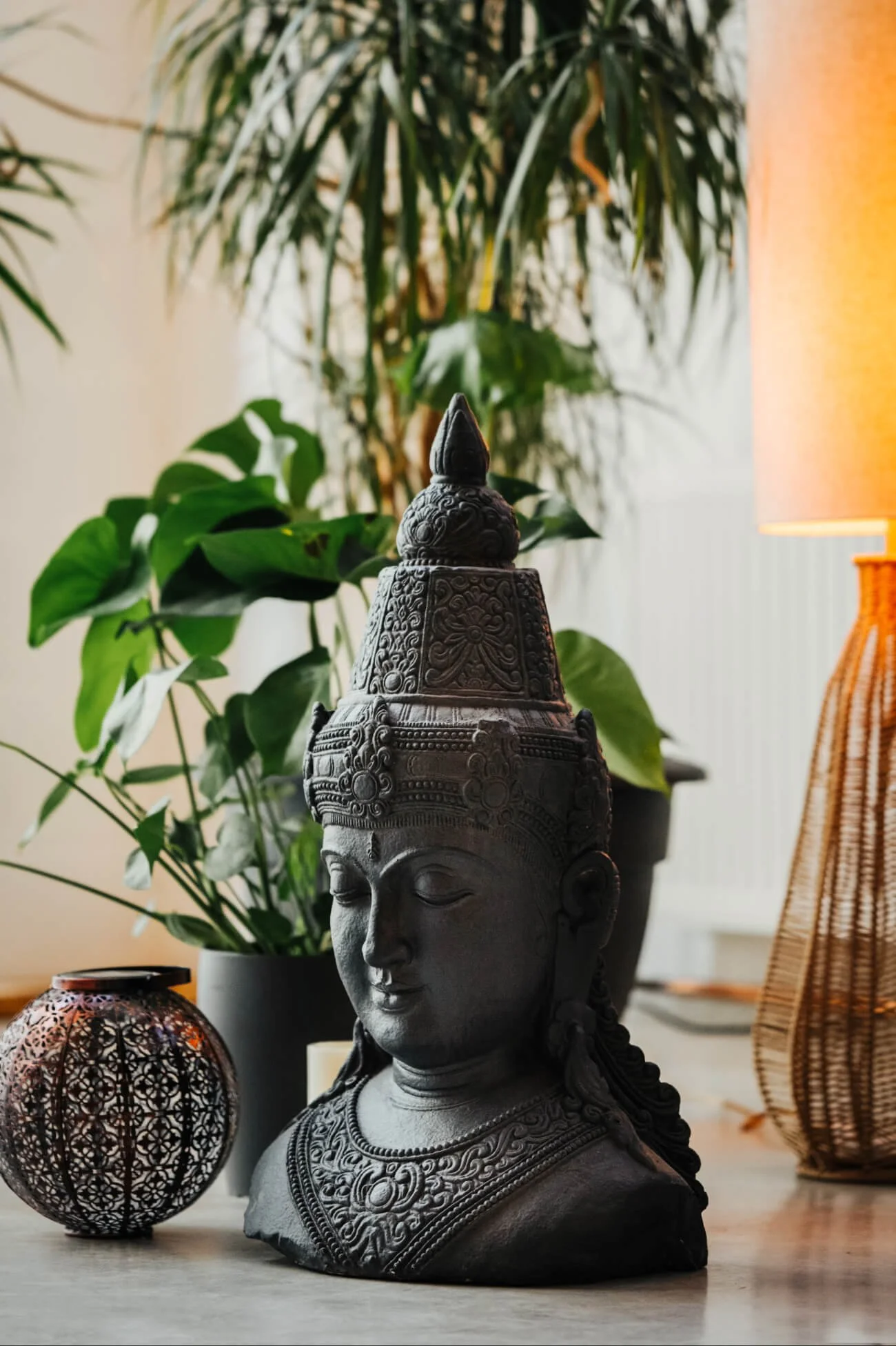 A black carved bust of a Buddha with detailed jewellery and headdress, placed on a cork flooring with decorative objects, against a backdrop of green plants and a lit lamp.