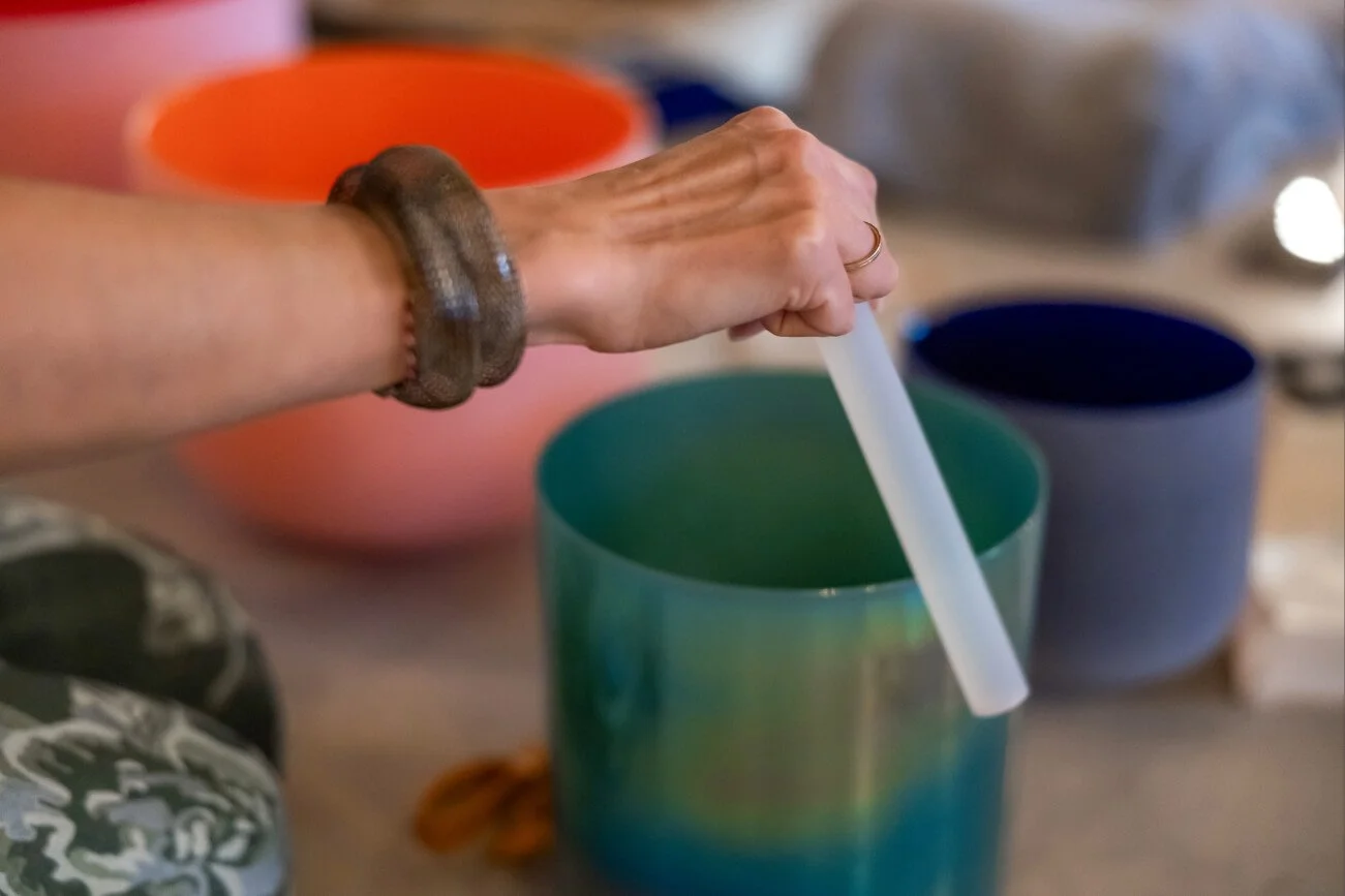 A person's hand holding a white stick above a green crystal sound bowl, with other colourful bowls in the background.