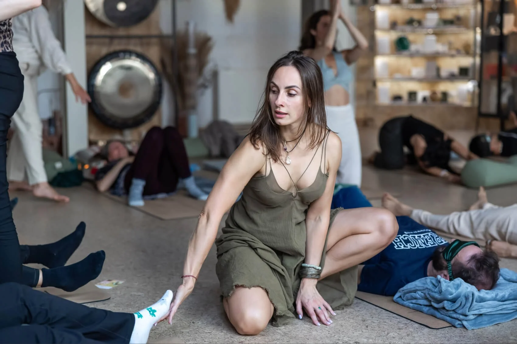 A woman in a green dress is kneeling on the floor, holding the foot of a person lying on the ground in a yoga class. Several other people are lying or sitting on mats around her, some in a similar relaxation pose. The setting looks like  wellness space.