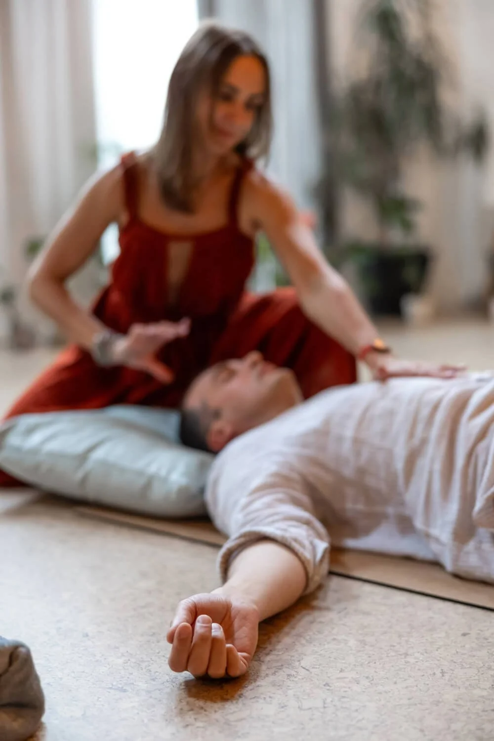 A woman is doing Reiki therapy on a person lying down on the floor with eyes closed, in a serene, well-lit room.
