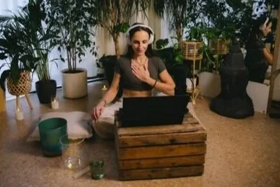 Woman sitting cross-legged on floor in front of laptop on a wooden table, wearing headphones and smiling.