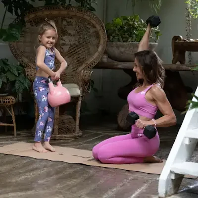 A woman in pink workout clothes doing strength training with dumbbells while kneeling on a yoga mat, smiling at a young girl holding a pink watering can in a lush, green indoor garden. Mat says LUV YOURSELF and shows a Kundalini snake.