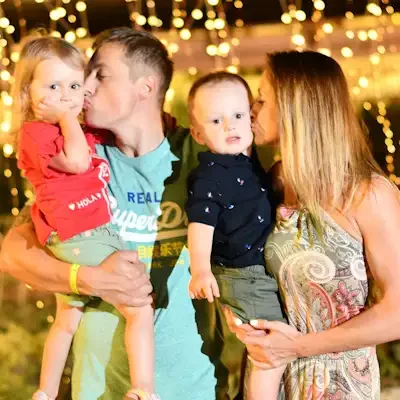 A family of four, two parents and two young children, sharing a kiss and holding them, during an outdoor evening event with string lights in the background.