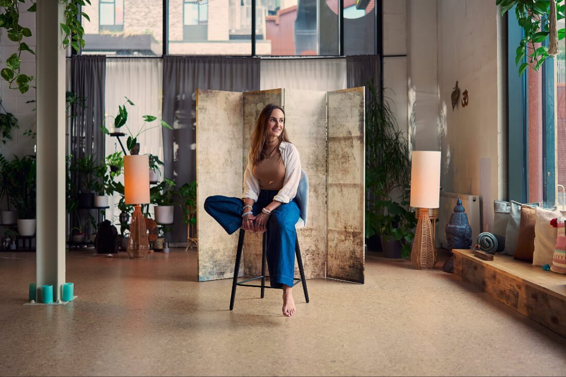 Woman sitting on a chair in a spacious, well-lit space with large windows, indoor plants, and decorative objects, smiling and looking to the side.
