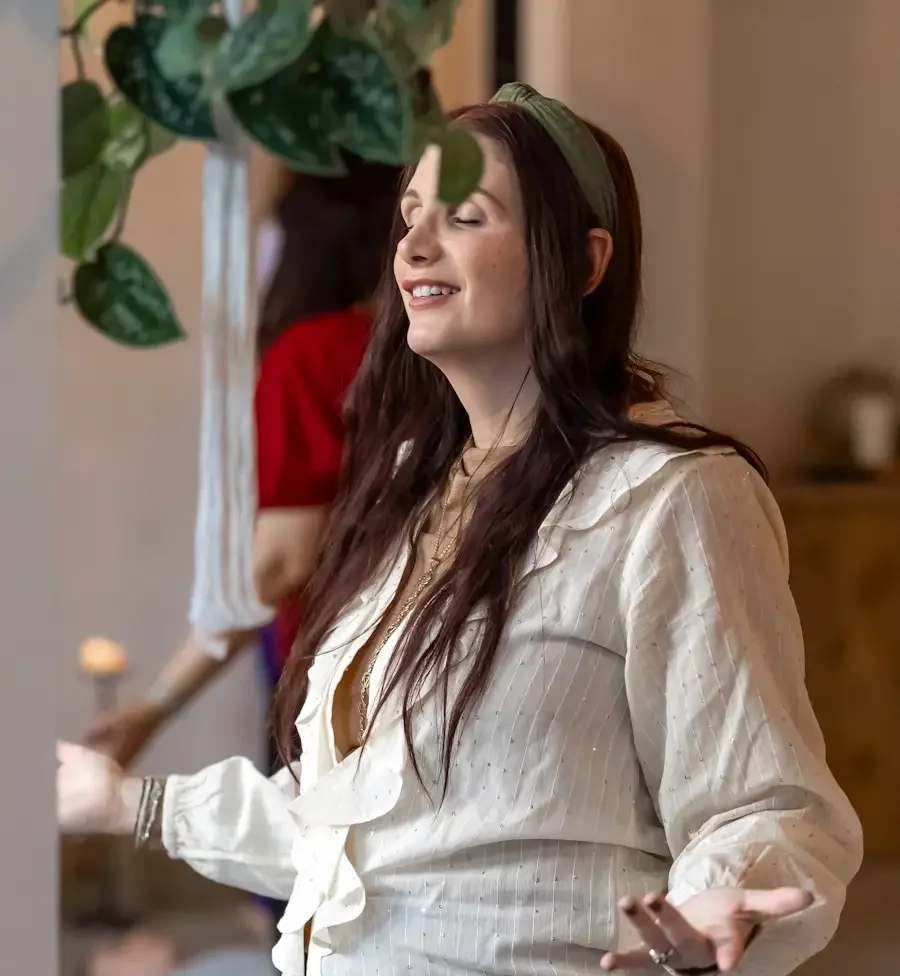 A woman with long dark hair and a headband smiling with eyes closed, wearing a cream-colored blouse with ruffles and brown pants, standing indoors with a plant in the foreground and another person blurred in the background.