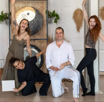 Five people posing and smiling in a bright, modern room with decorative plants and a yin-yang artwork on the wall. Holding certificates showing that they became Kundalini Activation Facilitators trained by Natalia Bareikis.