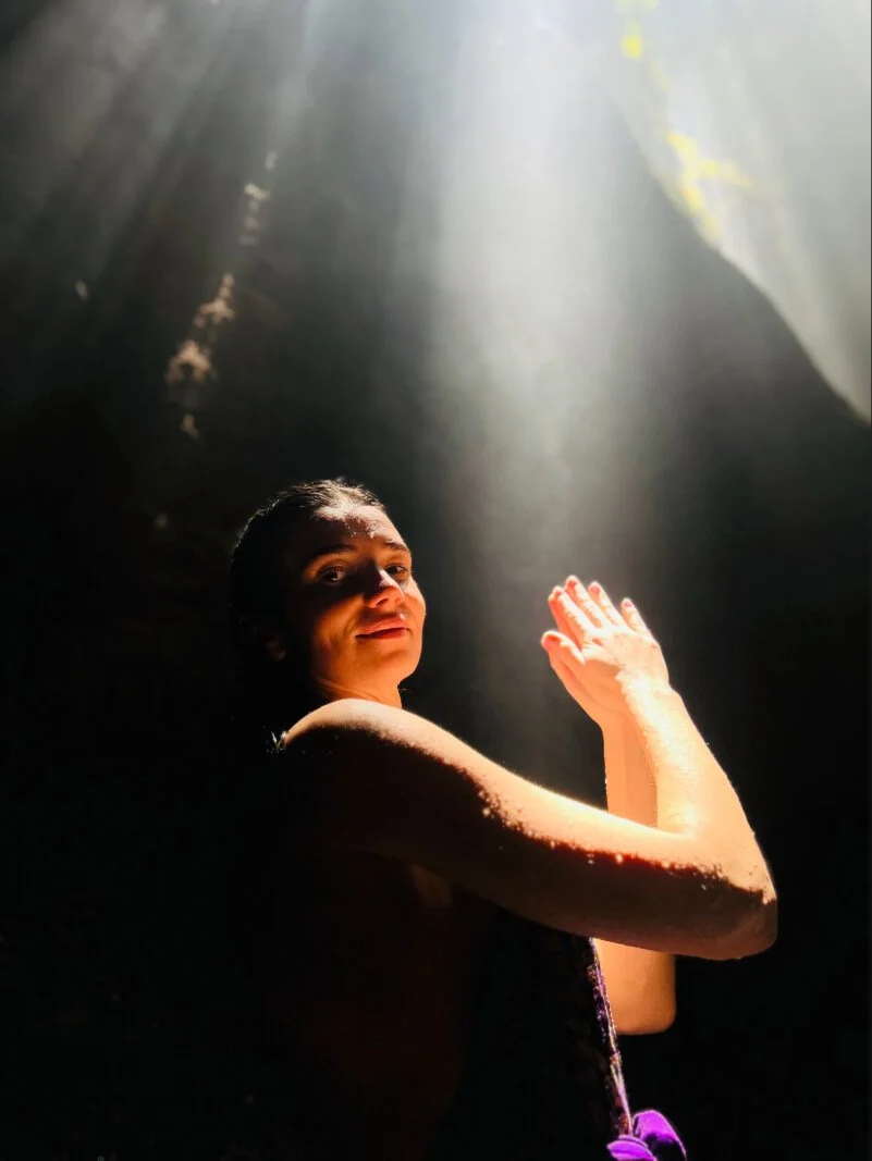 A woman with wet hair in a dark cave, illuminated by a beam of sunlight, turning her face and raised arm toward the light. Looks like a cave in Bali.