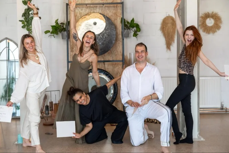 Five people posing and smiling in a bright, modern room with decorative plants and a yin-yang artwork on the wall. Holding certificates showing that they became Kundalini Activation Facilitators trained by Natalia Bareikis.