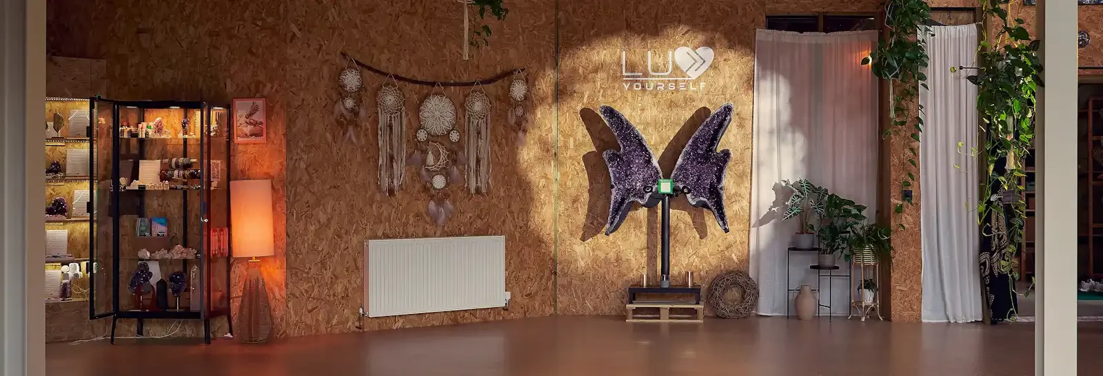 Decorative interior with a large butterfly sculpture, white curtains, potted plants, and hanging dreamcatchers on a wooden wall with the words 'LOVE YOURSELF' illuminated.