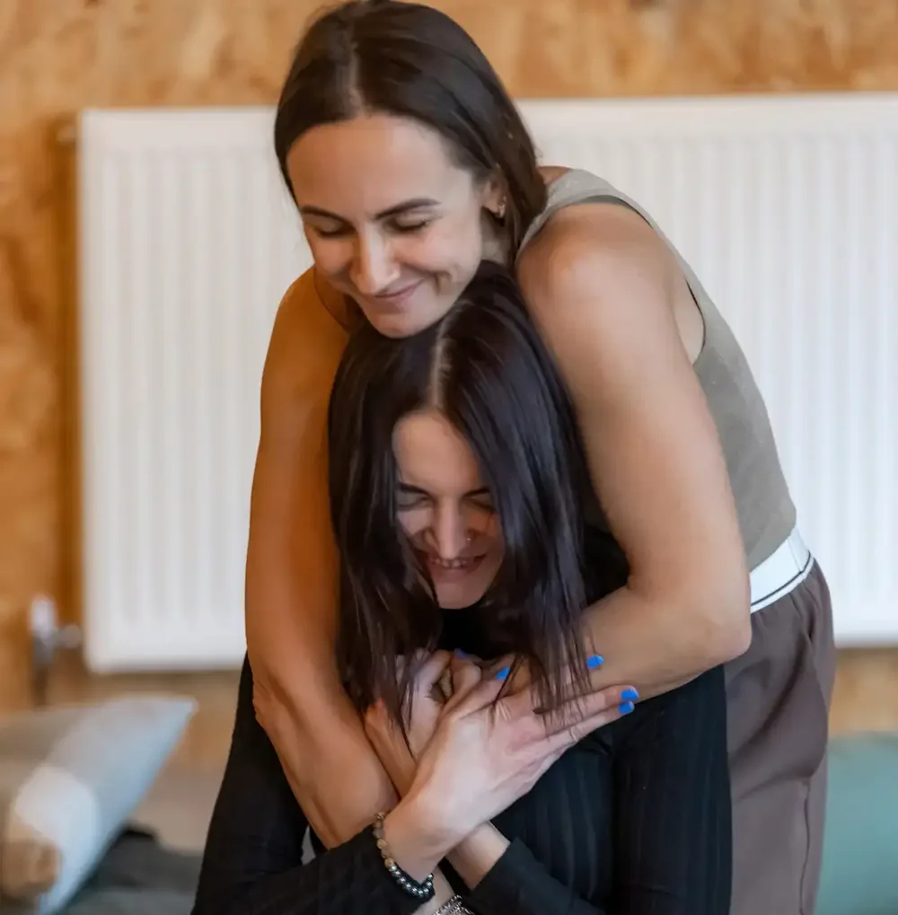 Two women hugging, smiling and sitting on the floor indoors with a wooden wall and a warm wooden wall in the background.