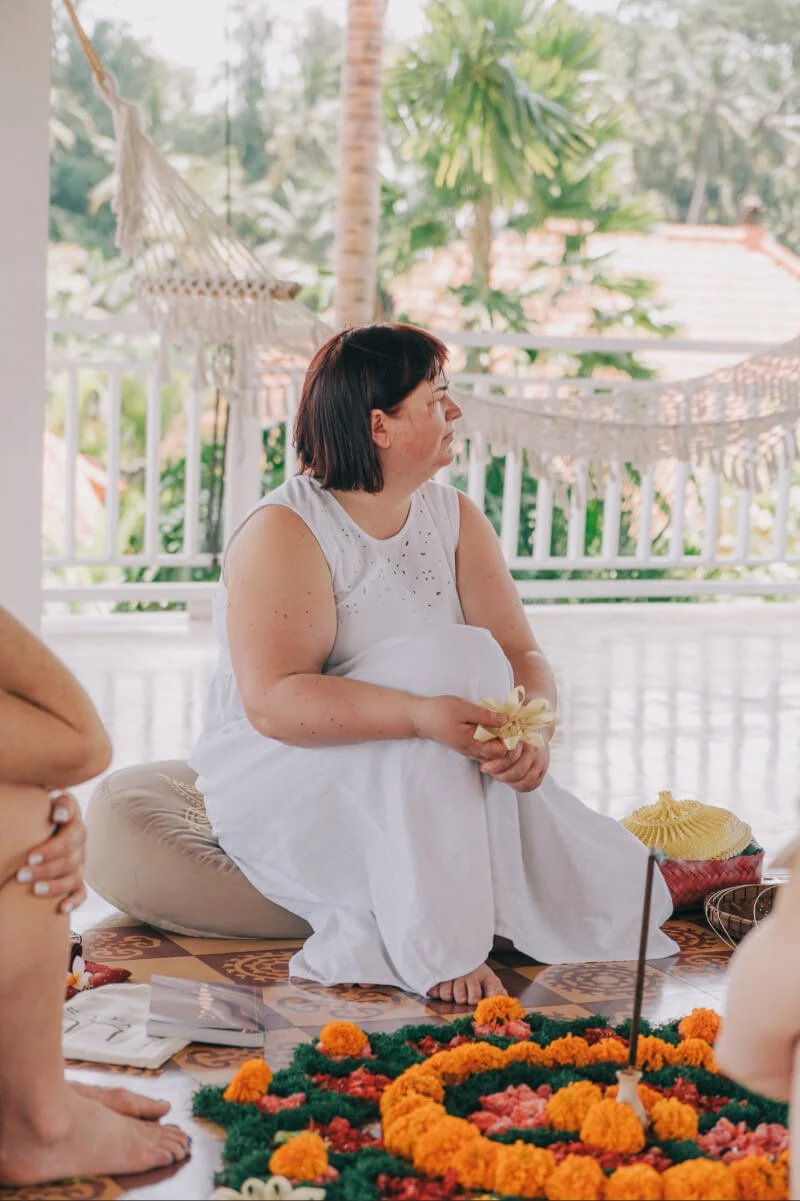A woman dressed in white sitting on the floor, holding a flower, during a Balinese ceremony outdoors with trees and plants in the background.