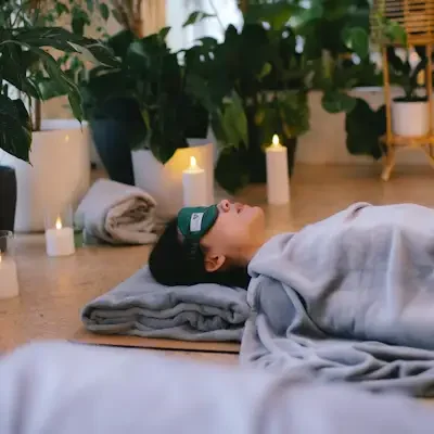 A person is lying on the floor with an eye mask on which says luv yourself by natalia and lady is covered with blanket with plants around her as well as candles.