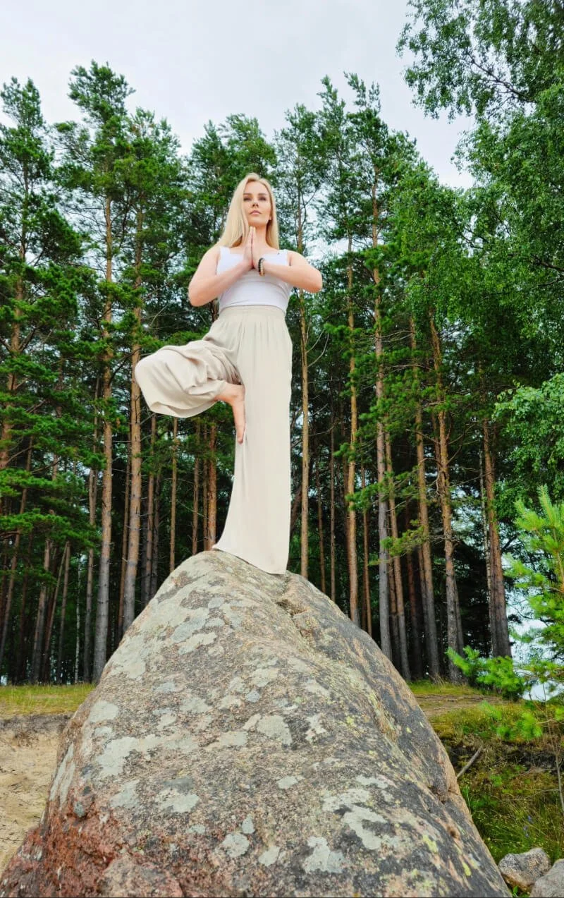 A woman practicing yoga outdoors standing on a large rock with one leg bent and hands in prayer position, surrounded by a forest of tall green trees.