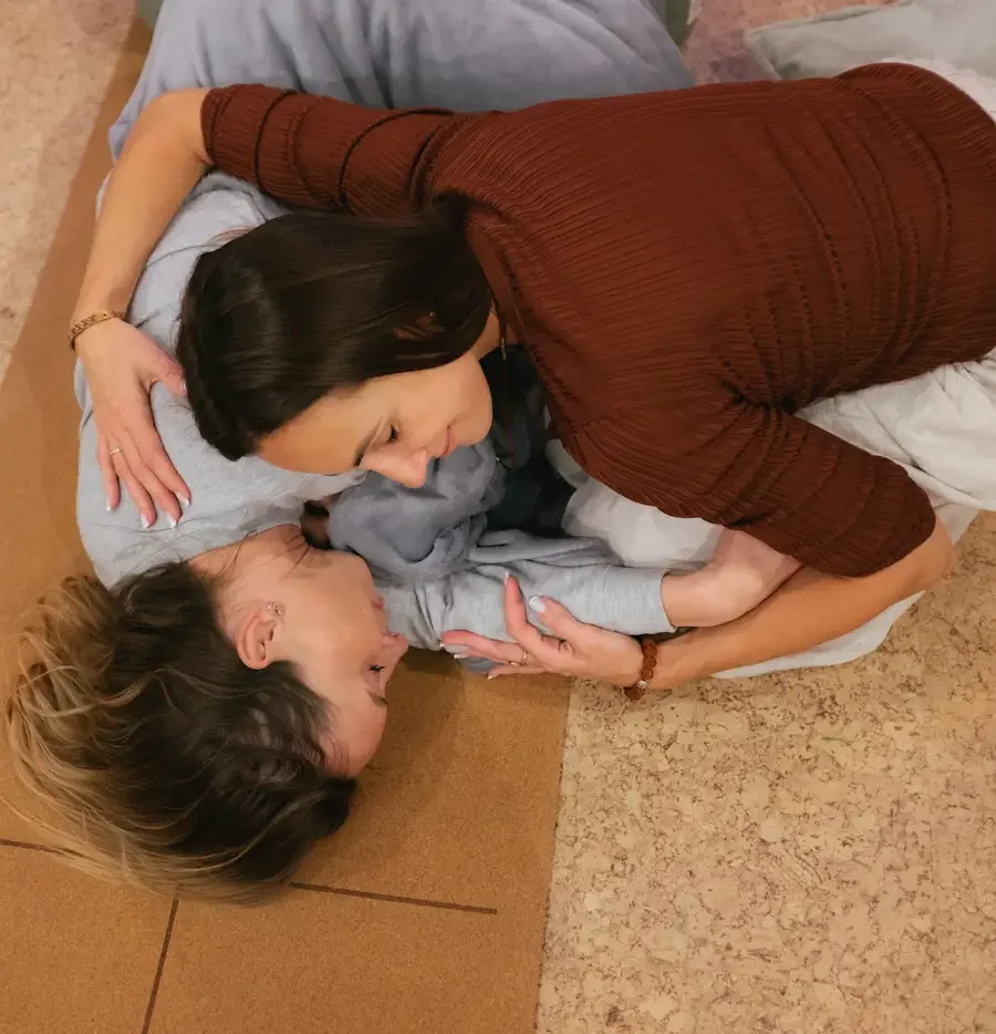 Two women on a brown mat, one assisting the other during childbirth in a medical setting.