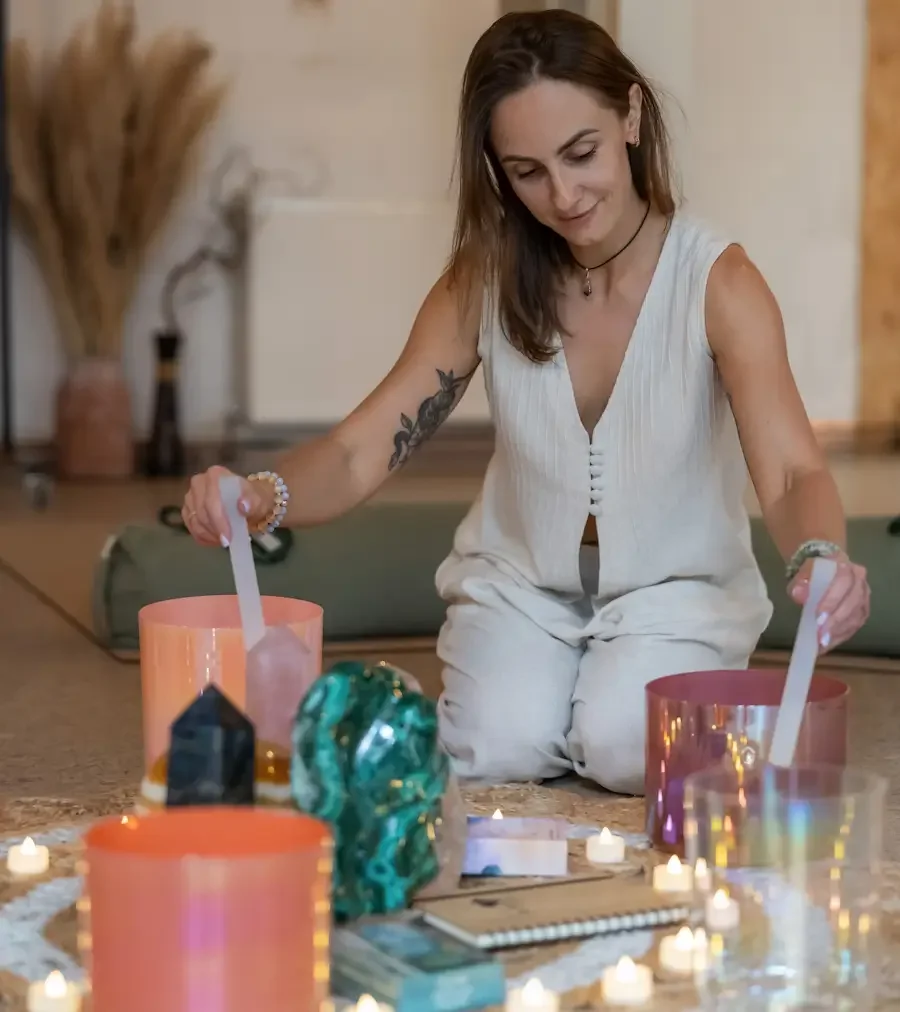 A woman with shoulder-length brown hair, snake tattoos on her right arms, and wearing a sleeveless white top is kneeling on the floor, surrounded by candles and crystals, performing a spiritual or meditative ritual in a serene space.