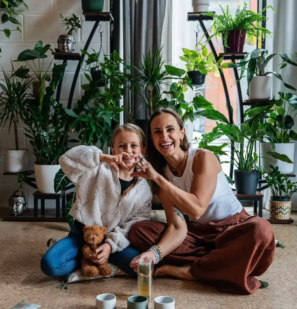 A woman is creating a heart with her fingers with a girl who looks like her daughter, sitting next to plants  and laughing