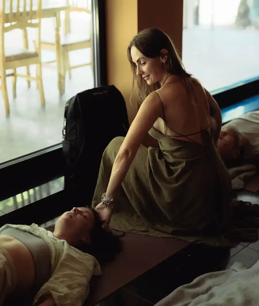 One woman is with a bliss on her face laying down on the floor and another woman is facilitating a session, another man is lying down on the other side with calm face