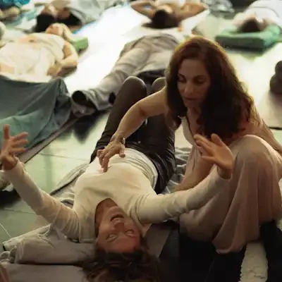 A woman and a girl doing healing session on a mat in a room with several other people lying down on mats behind them.