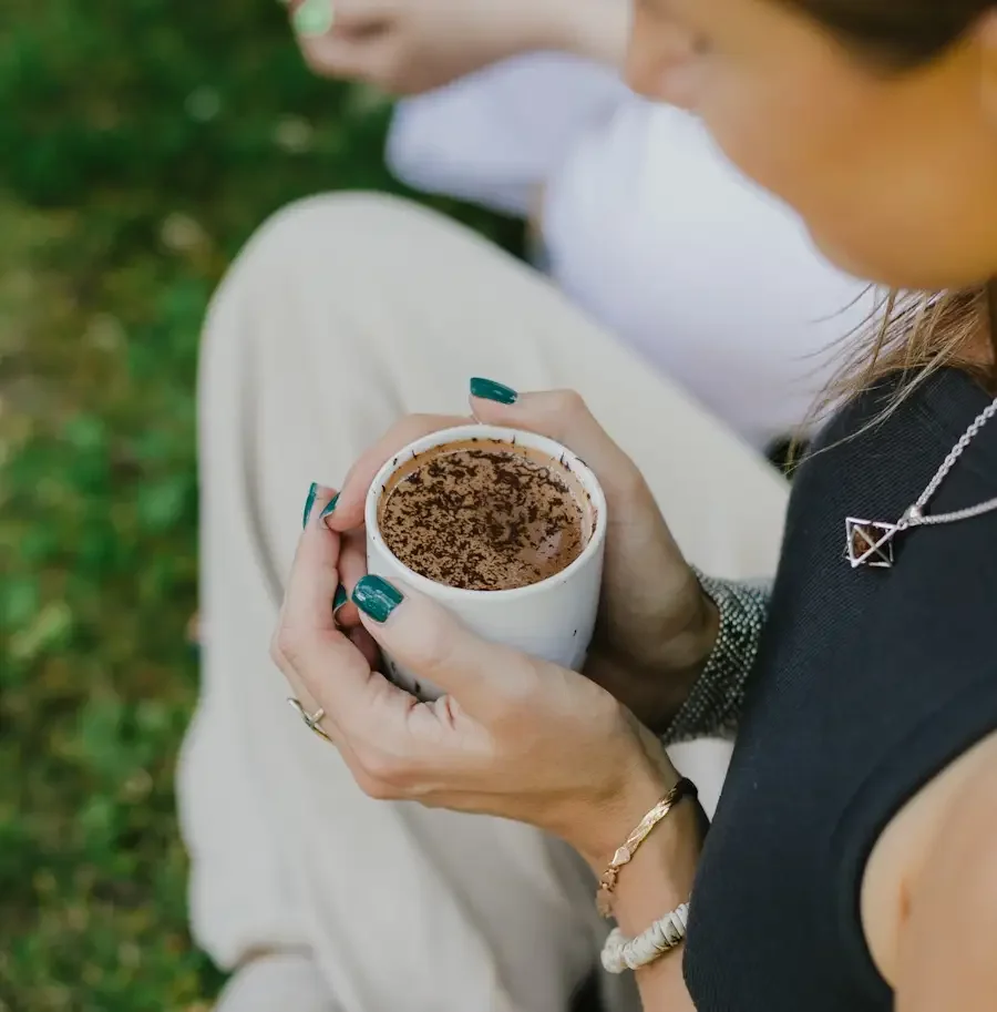 A woman is holding a mug of ceremonial cacao with chocolate shavings on top, sitting outdoors. Another person in the background is also holding a mug.