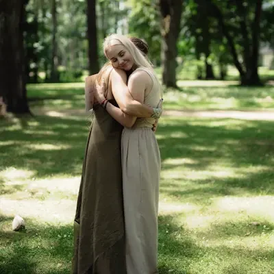 Two women embracing in the nature with tall trees and sunlight filtering through the leaves. They are calm and peaceful.