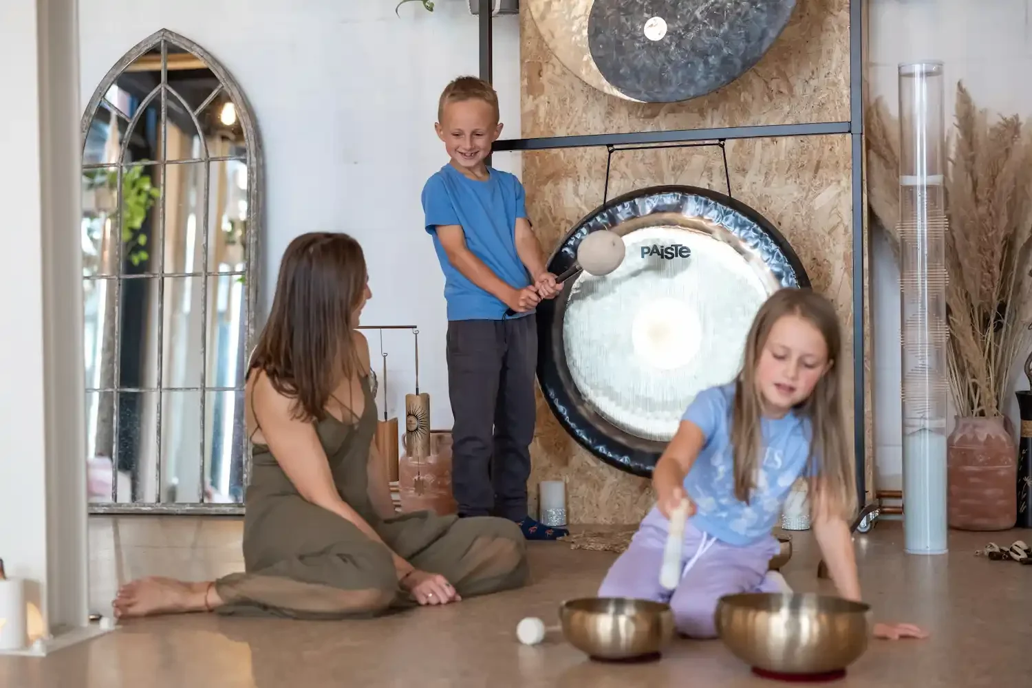 A woman on the floor in the serene space with two kids, all are smiling and playing boy a gong and girl is playing a tibetan bowl.