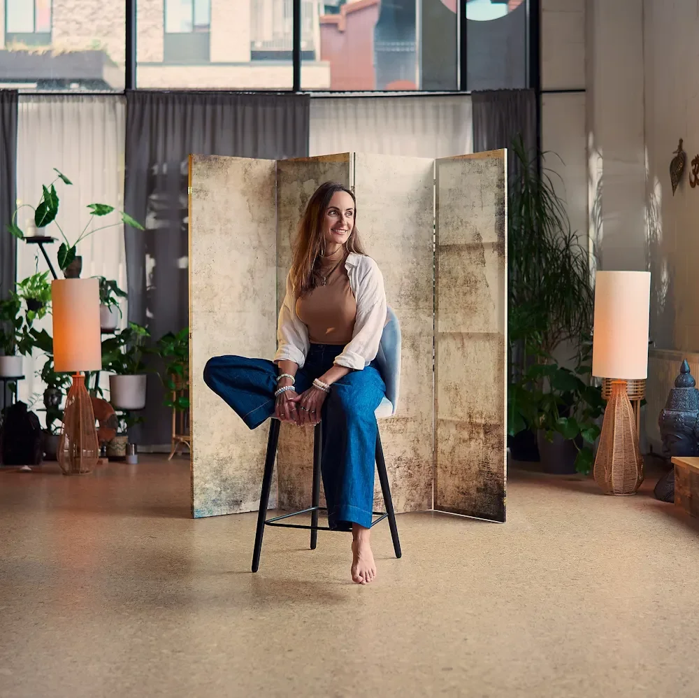 A woman sitting on a stool in a calm space with large windows, decorative room divider, plants, lamps, and cushions, looking to her right and smiling.