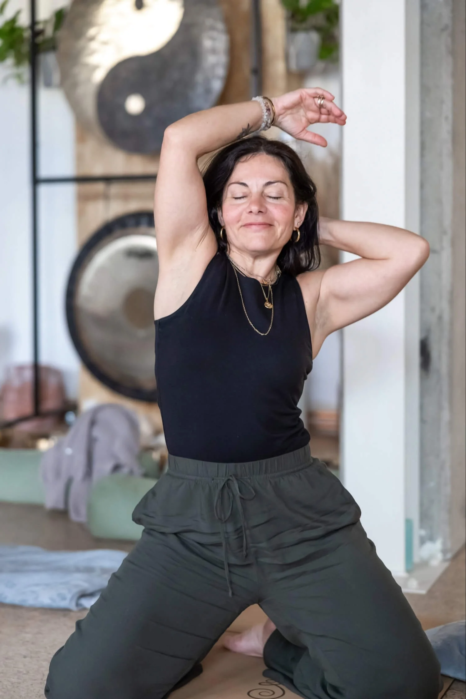 A woman in energetic flow and smiling, kneeling with one arm raised above her head and the other behind her neck, with a peaceful expression.