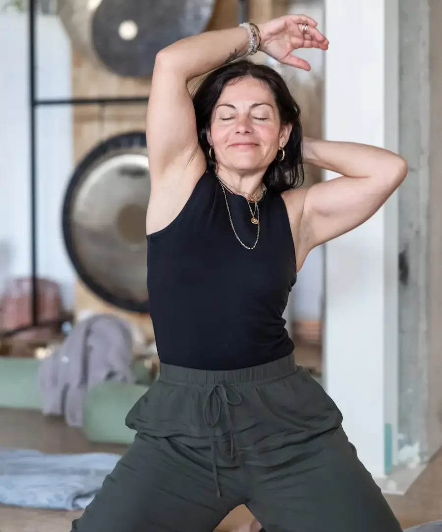 A woman in energetic flow and smiling, kneeling with one arm raised above her head and the other behind her neck, with a peaceful expression.