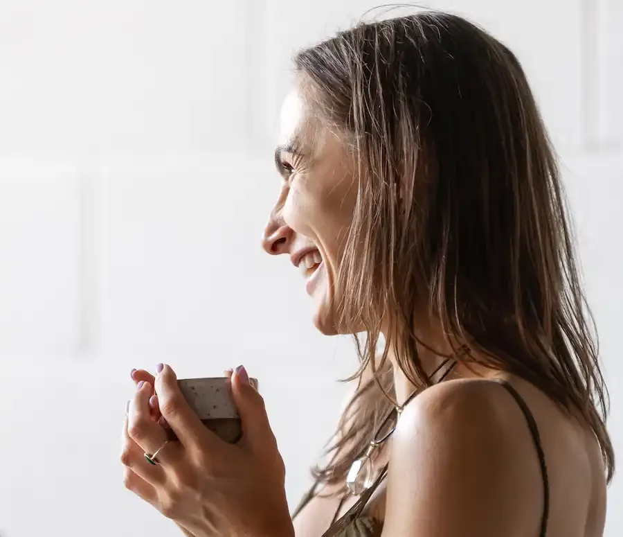 A woman with brown hair holding a cup with ceremonial cacao and smiling in a well-lit indoor setting.