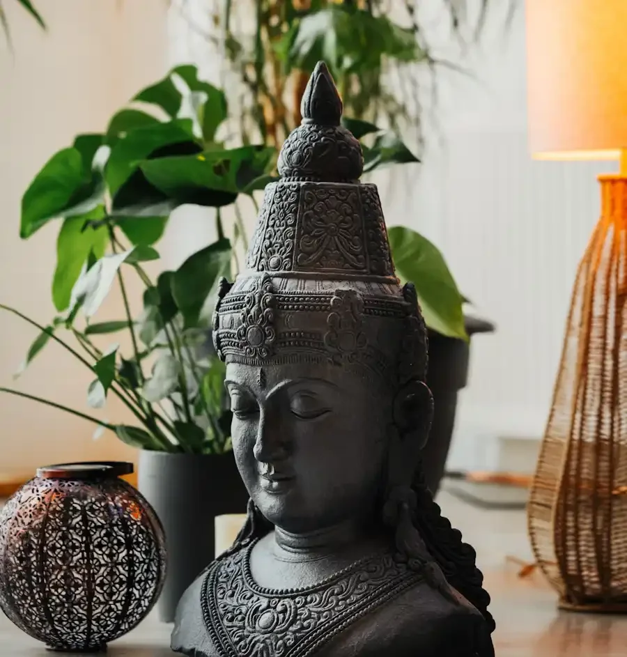 A detailed black sculpture of a Budda's bust, adorned with intricate jewellery and a tall, ornate crown, placed on a wooden surface with decorative lantern and plants in the background in a cosy space.