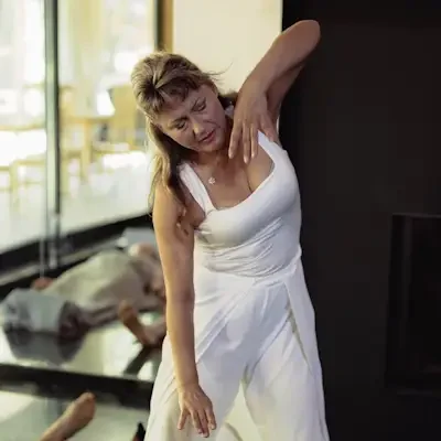 A woman in a white dress performs a healing session on a woman in a patterned dress lying on the floor in a living room with large windows and a fireplace.