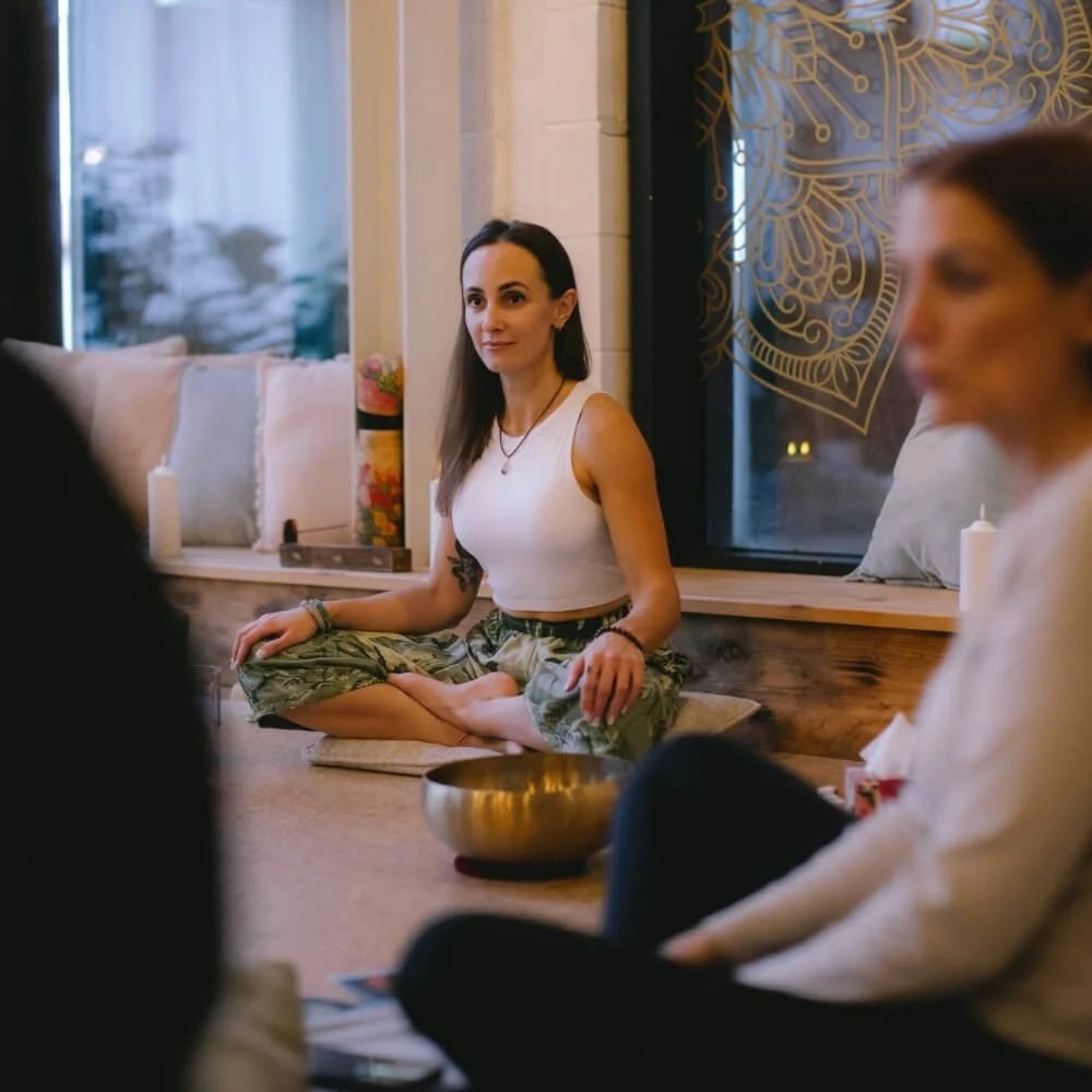 Woman sitting in lotus pose with a huambled face next to her a tibetan sound bowl and more people sitting on the floor.