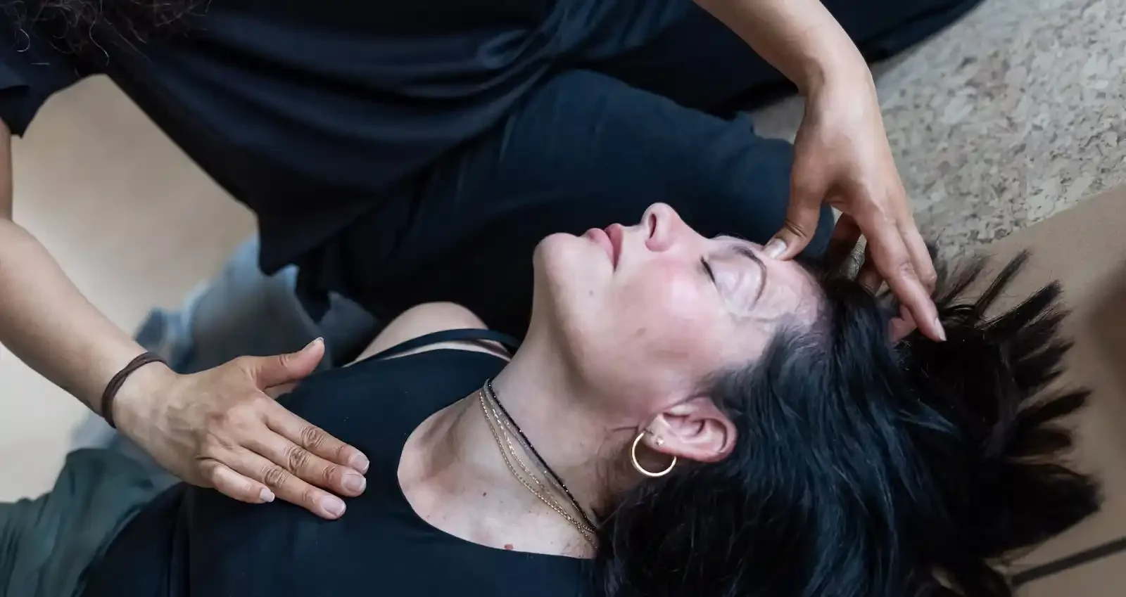 a woman peacefully on a cork yoga mat with a hand of another person on her heart and another hand is on her third eye