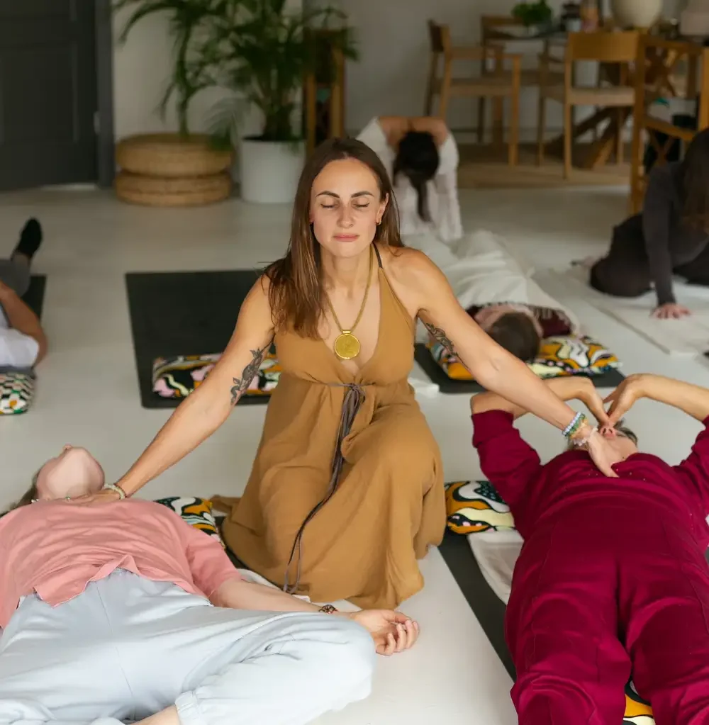 A woman facilitating the energy with her eyes closed while sitting on the floor, surrounded by people lying down or sitting with eyes closed in a cosy, well-lit room.