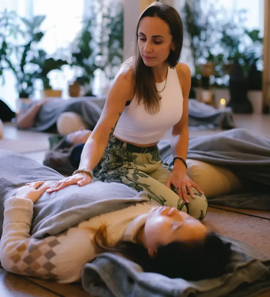 A woman giving a breathwork session to another person lying on a massage table in a cosy, well-lit room with plants in the background.