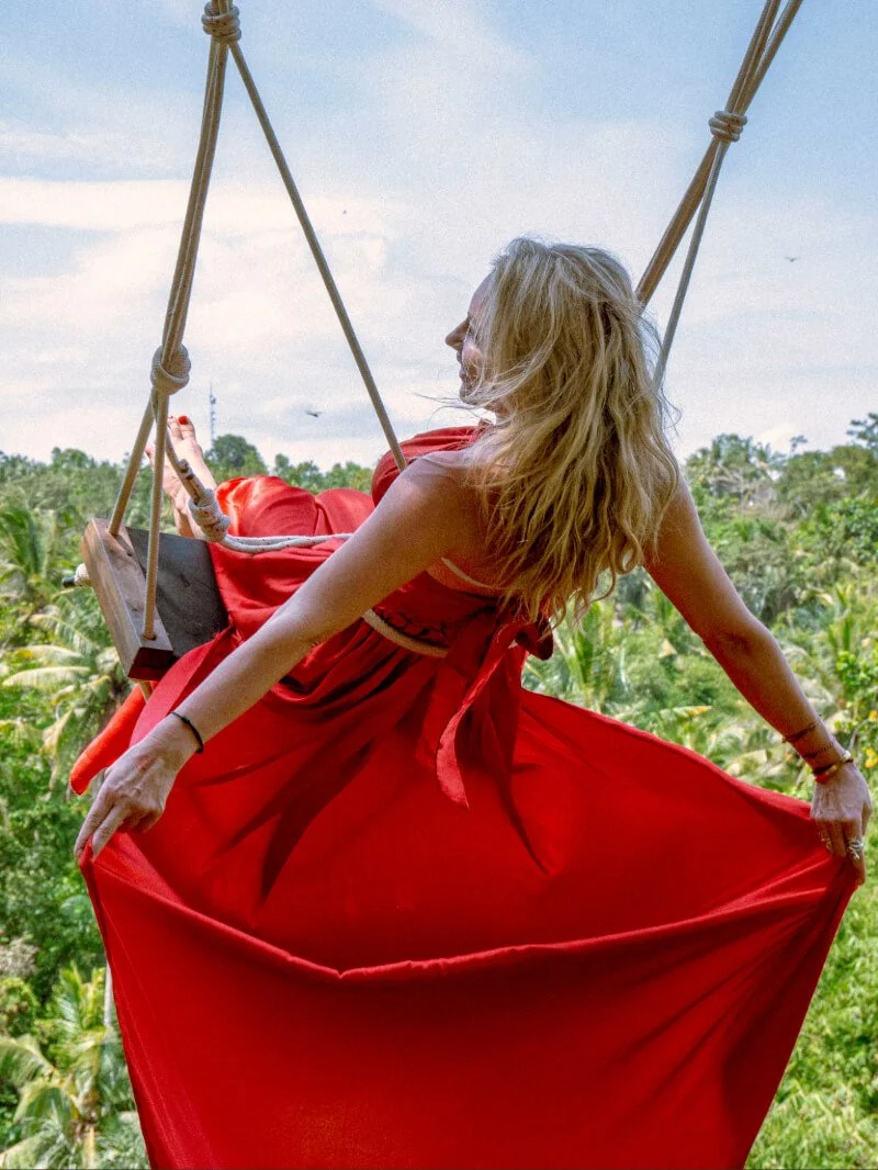 A woman with long blonde hair in a red dress sitting on a swing suspended in the air, overlooking a lush green forest and a blue sky with some clouds.