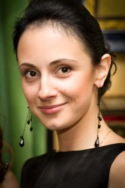 A young woman with dark hair in an updo, wearing earrings and a black top, smiling at the camera against a colorful background.