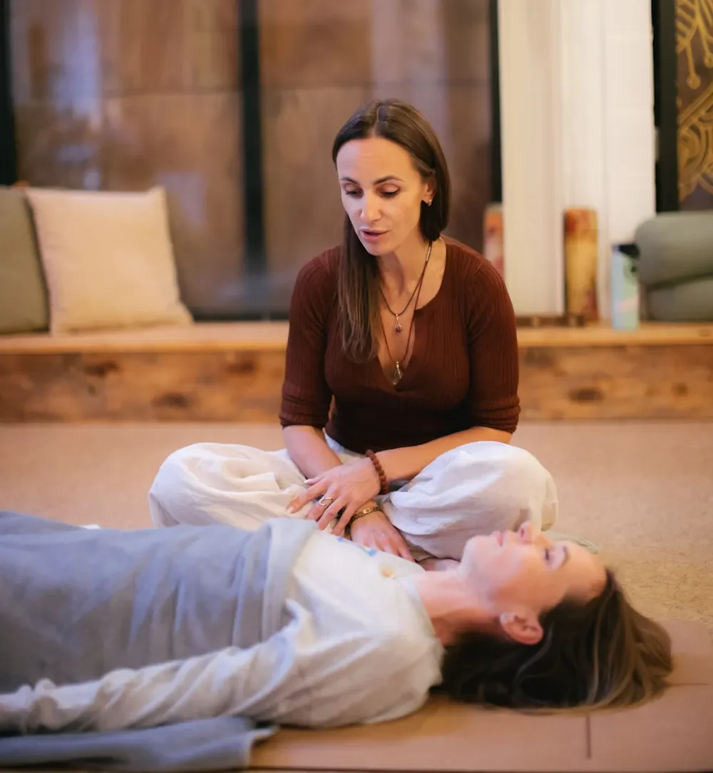 Two women practicing a guided breathwork or energy healing session in a cosy, warmly lit room. One woman lies on the floor with her eyes closed, while the other sits beside her, appearing focused and attentive.