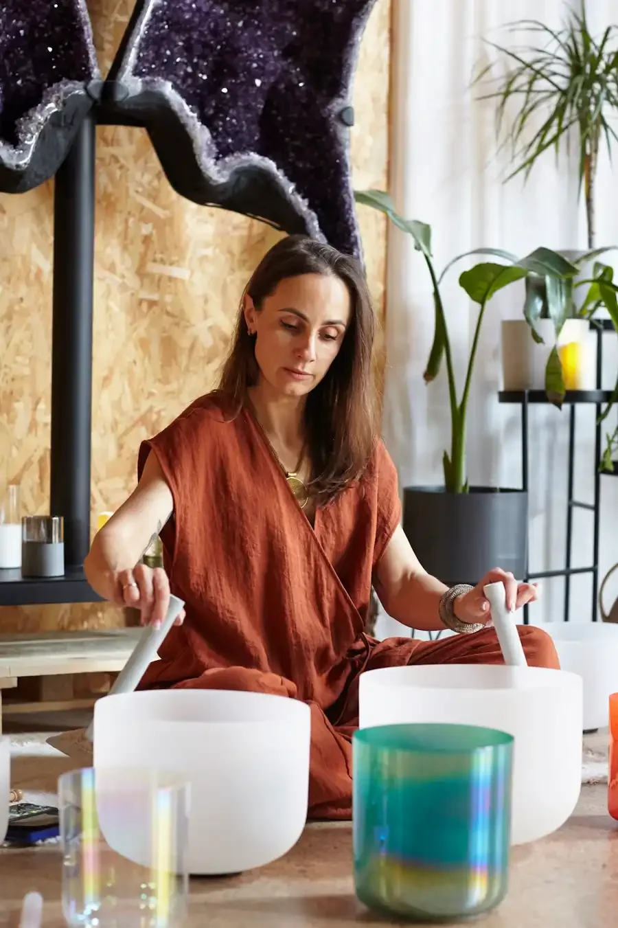 A woman sitting on the floor playing crystal bowls with candles and plants around and large crystal behind her