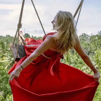 A woman with long blonde hair in a red dress sitting on a swing suspended in the air, overlooking a lush green forest and a blue sky with some clouds.