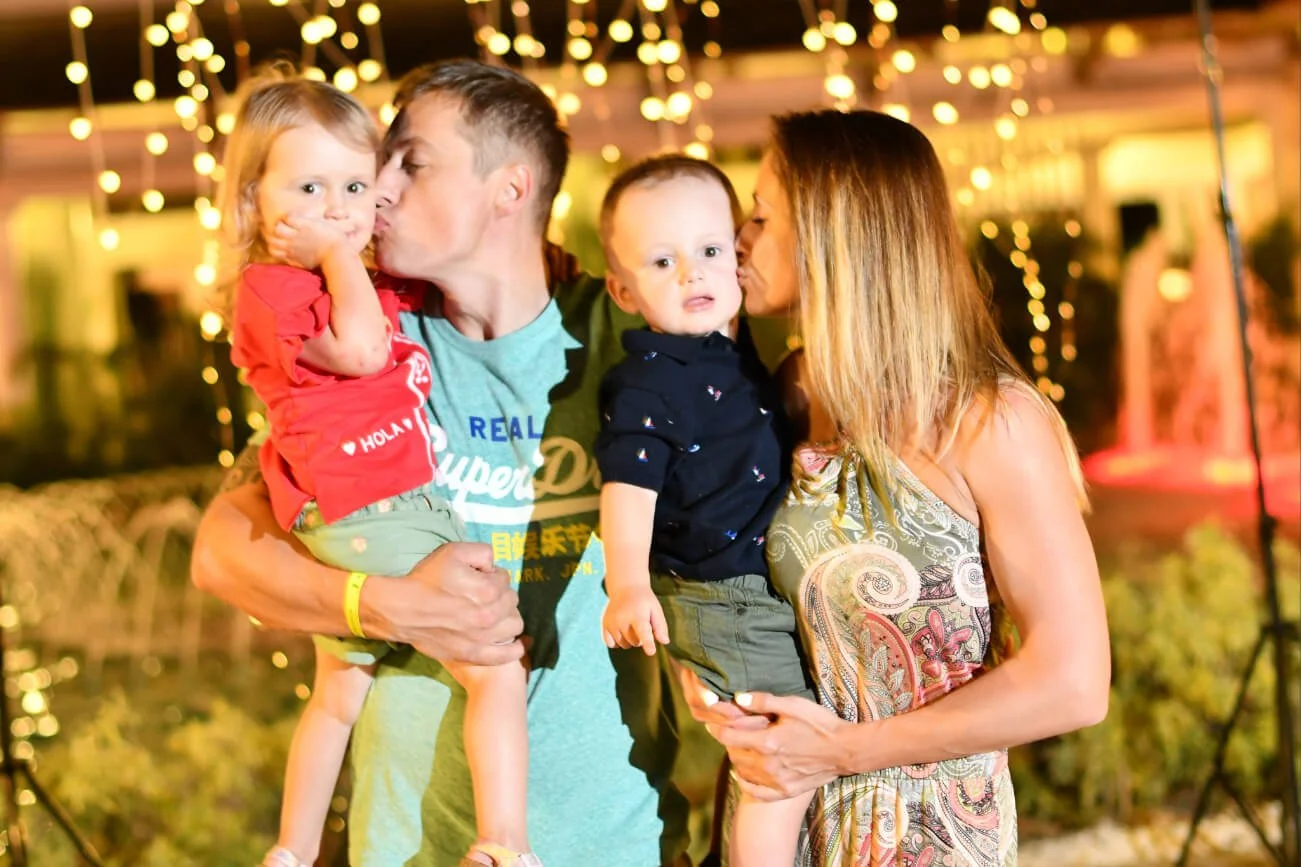 A family of four, two parents and two young children, sharing a kiss and holding them, during an outdoor evening event with string lights in the background.