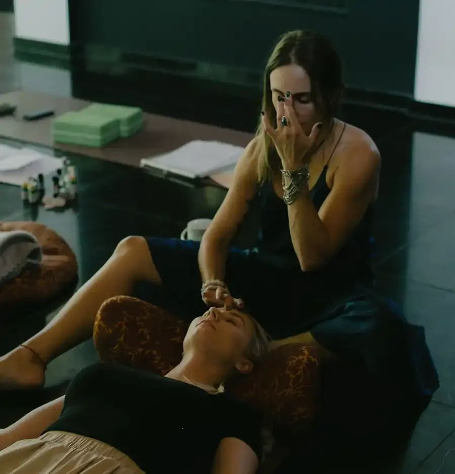 Two women, one lying down wearing beige pants and black top with her eyes closed, the other sitting on the floor with hands covering her face, giving a facial treatment or massage in a dimly lit room.