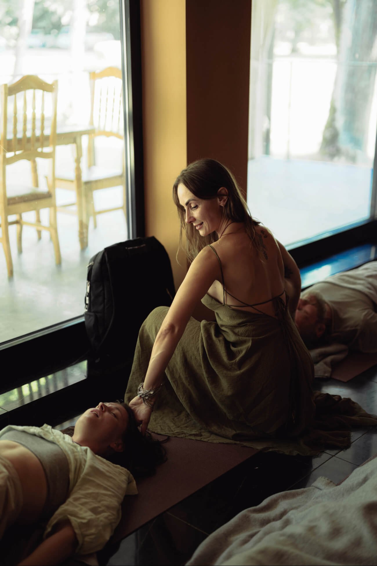 A woman sitting on her knees, with a smile, comforting another woman lying on the floor with her eyes closed, near a large window with chairs outside.