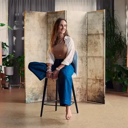 Woman sitting on a chair in a spacious, well-lit space with large windows, indoor plants, and decorative objects, smiling and looking to the side.