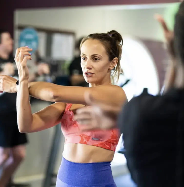 A woman in a pink sports bra and purple leggings is stretching her arm in a gym, with other people exercising in the background.