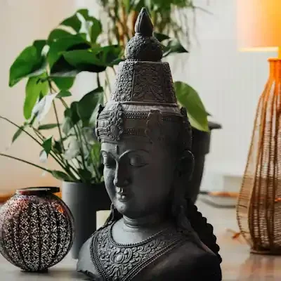 A black carved bust of a Buddha with detailed jewellery and headdress, placed on a cork flooring with decorative objects, against a backdrop of green plants and a lit lamp.