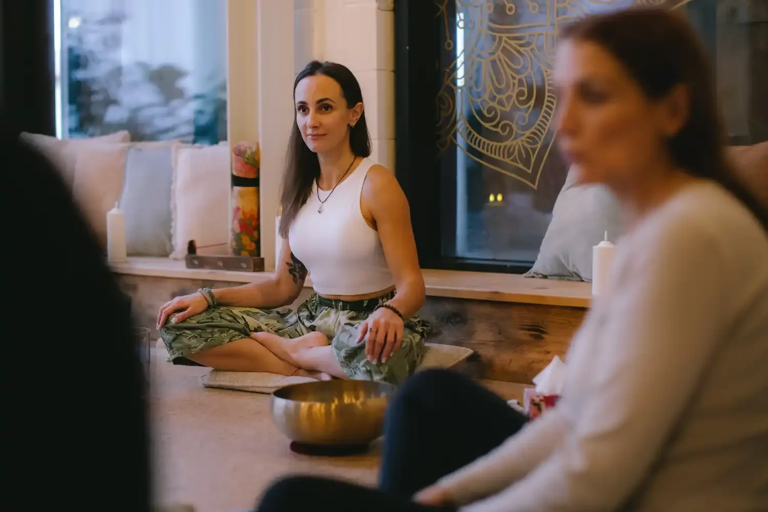 Woman sitting in lotus pose with a humbled face next to her a tibetan sound bowl and more people sitting on the floor.