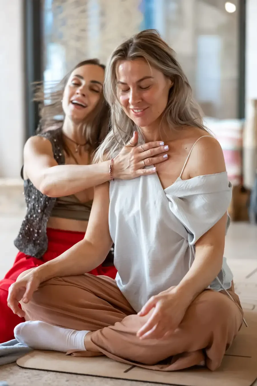 Two women practicing energy healing, with one woman placing her hand on the chest of the other woman who is sitting cross-legged on the floor, eyes closed, smiling, in a brightly lit indoor setting.