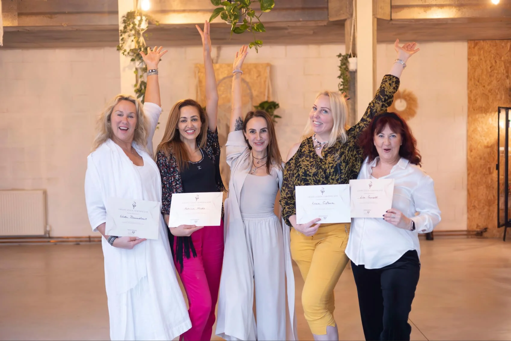 Five women standing together, some holding certificates, celebrating at an indoor event, with their arms raised and smiling. Finishing Kundalini Activation facilitator training with Natalia Bareikis as it says on certificate.